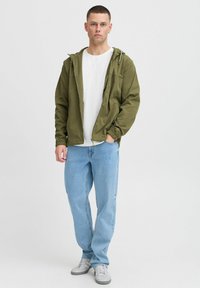 Jachetă ușoară de culoare verde măslinie cu glugă, cu închidere cu fermoar și buzunare, purtată peste un tricou alb și blugi bleumarin. Adidașii completează ținuta.