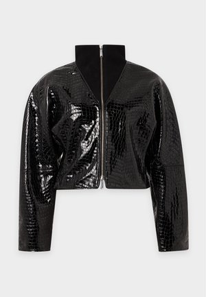 Jeune femme portant une veste noire texturée, un haut blanc, une jupe pied-de-poule, des bottes noires décontractées, tenant un sac à main noir, debout sur fond blanc.