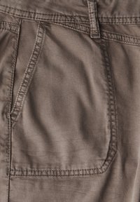 Nära bild av brunt tyg som visar en framficka med synliga sömnadsdetaljer. Materialet har en slät, något texturerad yta.