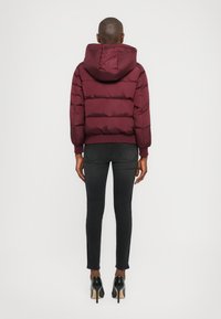 Burgunderfarget boblejakke med hette, med horisontale quiltedetall, kombinert med tettsittende svarte jeans og svarte høye hæler.