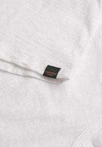 Witte katoenen t-shirt met een gladde textuur, met een zwart label dat aan de zoom is gestikt met oranje tekst die "Superdry" leest.