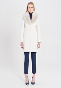Cappotto bianco in lana con colletto in pelliccia sintetica, caratterizzato da un taglio dritto, cinque bottoni, abbinato a jeans in denim scuro e tacchi blu.