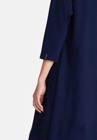 Vestido azul marino con mangas de tres cuartos, textura suave, forma ligeramente acampanada, que muestra una pequeña etiqueta de logo en el puño.