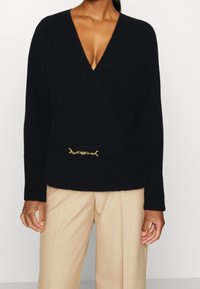 Suéter negro de envoltura con un profundo escote en V, mangas largas, una textura suave y un detalle de cadena dorada en la cintura. Combinado con pantalones beige.