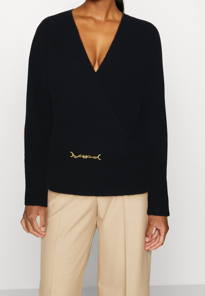 Suéter negro de envoltura con un profundo escote en V, mangas largas, una textura suave y un detalle de cadena dorada en la cintura. Combinado con pantalones beige.