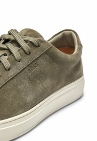 Green suede sneaker with rounded toe, flat white sole, and tonal laces. Features embossed brand logo on the side and soft inner lining.