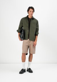 Camisa de botón verde oscuro sobre un suéter marrón, pantalones cortos a rayas beige, calcetines blancos y zapatos negros. Sosteniendo una bolsa negra. Fondo simple.