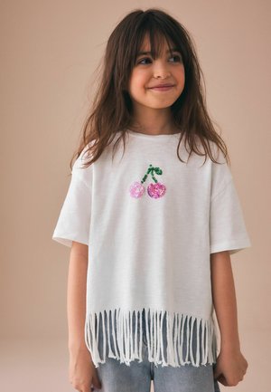 Hosszú hajú fiatal lány, fehér pólót viselve, rózsaszín cseresznye mintával és rojtos szegéllyel, bézs háttér előtt áll.