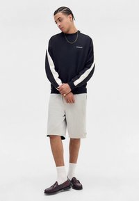 Čierna mikina s dlhým rukávom s bielymi bočnými panelmi, skombinovaná so sivými perforovanými šortkami. Oblečenie dopĺňajú hnedé mokasíny a biele ponožky nad členky.