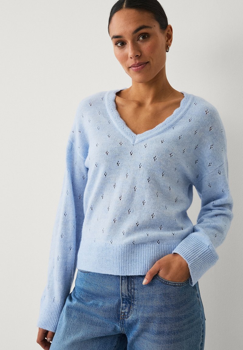 Žena s tmavými vlasy v světle modrém svetru s výstřihem do V a vzorem drobných otvorů, oblečená v modrých džínách, pózující s jednou rukou v kapse.