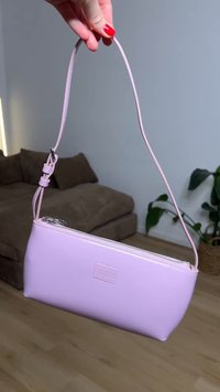 Main avec du vernis à ongles rouge tenant un petit sac à main violet clair par sa bandoulière à l'intérieur près d'un canapé et d'une plante.