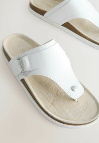 Sandales en cuir blanc à lanière ajustable, semelle intérieure en suède beige rembourrée et semelle extérieure en caoutchouc blanc sur fond clair.