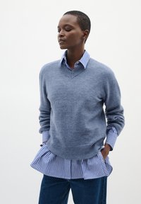 Siv pleteni pulover s V-izrezom, nosen čez modro-belo črtasto srajco, s nabranim robom in modrimi hlačami visokega pasu.