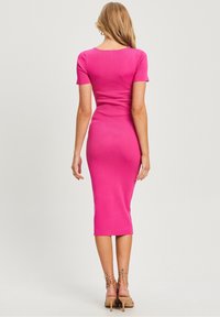 Abito rosa aderente con maniche corte, orlo a ginocchio e texture a coste. Indossato con sandali con tacco nudo e cinturini.