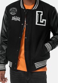 Osoba w czarnej kurtce varsity z skórzanymi rękawami i logo Lonsdale, w pomarańczowej koszulce, stojąca z jedną ręką w kieszeni.