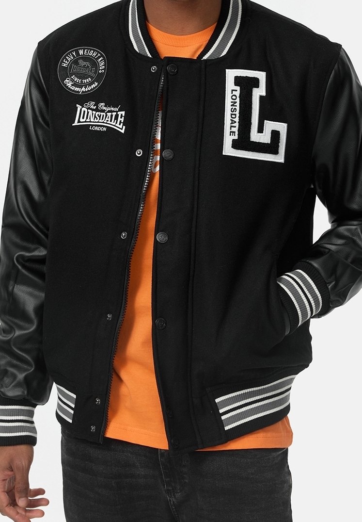 Osoba w czarnej kurtce varsity z skórzanymi rękawami i logo Lonsdale, w pomarańczowej koszulce, stojąca z jedną ręką w kieszeni.