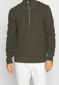 Dunkelgrüner gestrickter Pullover mit hohem Kragen und halbem Reißverschluss. Besitzt eine gerippte Textur und lange Ärmel, kombiniert mit weißen Hosen.