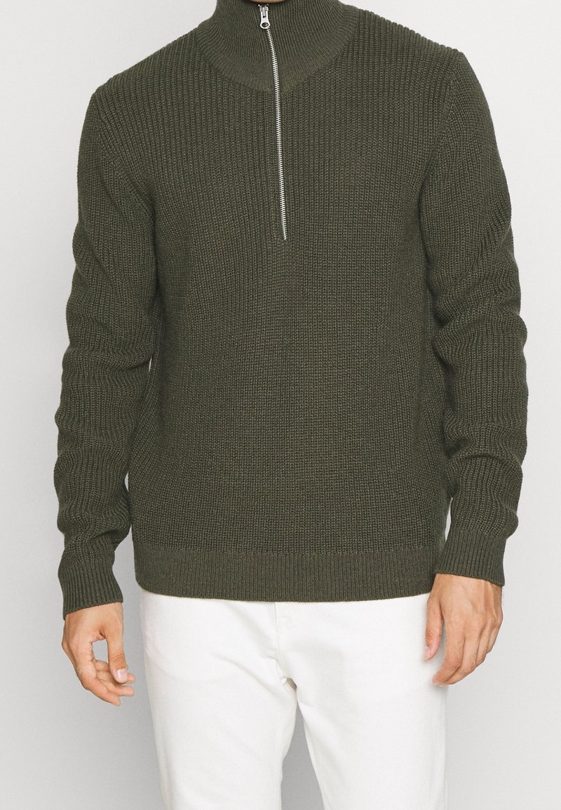 Dunkelgrüner gestrickter Pullover mit hohem Kragen und halbem Reißverschluss. Besitzt eine gerippte Textur und lange Ärmel, kombiniert mit weißen Hosen.