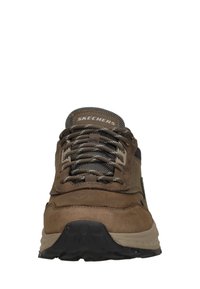 Zapatilla de ante marrón con una parte superior texturizada, que presenta acentos oscuros y cordones grises. Suela de goma gruesa con un patrón de dibujo. Logo "SKECHERS".