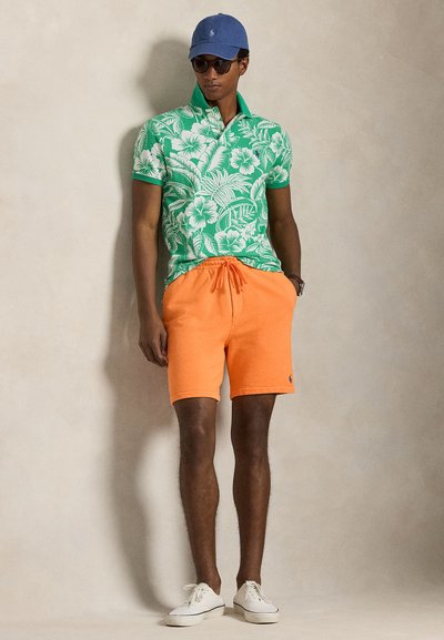 Άνδρας με πράσινο φλοράλ πόλο, φωτεινά πορτοκαλί σορτς, μπλε καπέλο, γυαλιά ηλίου και λευκά αθλητικά παπούτσια στέκεται μπροστά σε απλό φόντο.