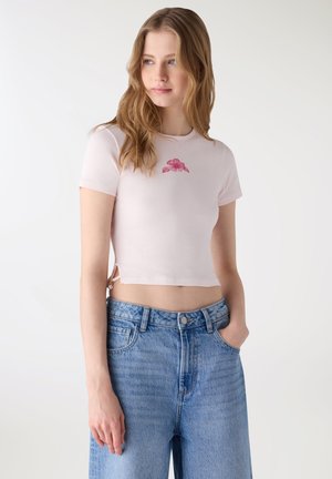 Giovane donna che indossa una T-shirt corta rosa pallido con un piccolo disegno di fiore rosa, abbinata a jeans blu a vita alta, in piedi con una mano in tasca.
