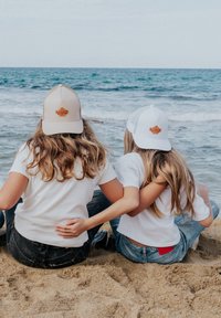 Gorras de camionero en beige y blanco con parches de cuero, combinadas con camisetas blancas y jeans de mezclilla, sentadas en una playa de arena junto al océano.