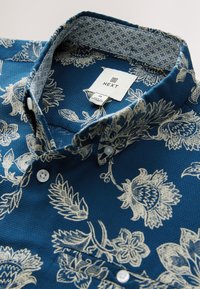 Camicia blu a fantasia con disegni floreali color crema. Tessuto strutturato con bottoni e colletto interno a contrasto. Etichetta del brand visibile.