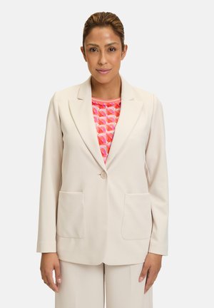 Hellbeige Blazer mit einem einzelnen Knopf, Reverskragen und zwei vorderen Taschen; getragen über einem pinkfarbenen geometrisch gemusterten Oberteil. Glattes Material.