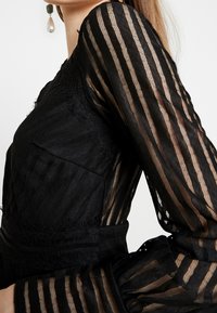 Černý oděv s průhlednými, proužkovanými dlouhými rukávy, texturovanou tkaninou a řasenými akcenty na manžetách, předvádějící složité detaily a design.