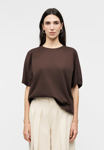 Mujer con cabello largo y oscuro, que lleva pendientes de aro dorados, una blusa de manga corta suelta de color marrón oscuro y pantalones beige de talle alto con pliegues.