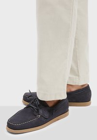 Marineblaue Wildlederschuhe mit flacher Gummisohle, dekorativem Nähten und einer Bindung; kombiniert mit hellbeigen Hosen.