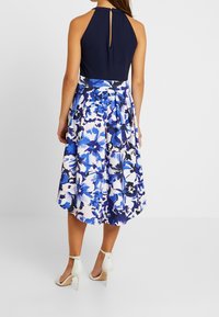 Kwiatowa sukienka z granatową górą na halter i niebieską spódnicą w kwiatowy wzór. Posiada dekolt w kształcie łezki, zwiewny fason oraz białe sandały na obcasie.