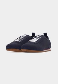 Paire de baskets décontractées bleu marine avec des lacets blancs, des semelles en caoutchouc marron et des languettes au talon sur un fond clair uni.