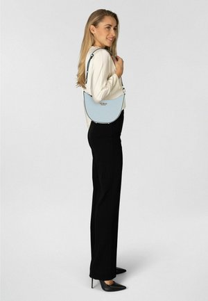 Donna che indossa pantaloni neri, top beige, tacchi neri, con una borsa Guess azzurro chiaro a tracolla, in piedi di lato e sorridente.