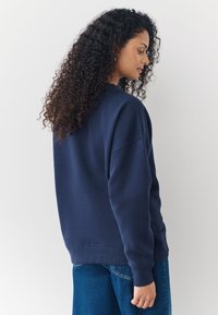 Tamnoplavi oversized duks s rebrastim manžetnama i rubom. Mekani materijal glatke teksture, s opuštenim krojem i spuštenim ramenima.