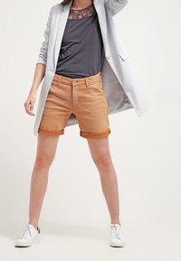 Shorts en denim marron clair avec des revers, assortis à un haut gris et une légère veste, complétés par des baskets blanches.