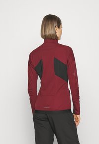 Bordeauxfarbene Sportjacke mit schwarzen Akzenten, hohem Kragen und tailliertem Design. Strukturierter Stoff mit reflektierenden Details auf der Rückseite.