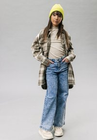 Bruine geruite shirtjacket over een beige top, blauwe wijde jeans en crèmekleurige sneakers. Felgele gebreide beanie. Grauwe achtergrond.