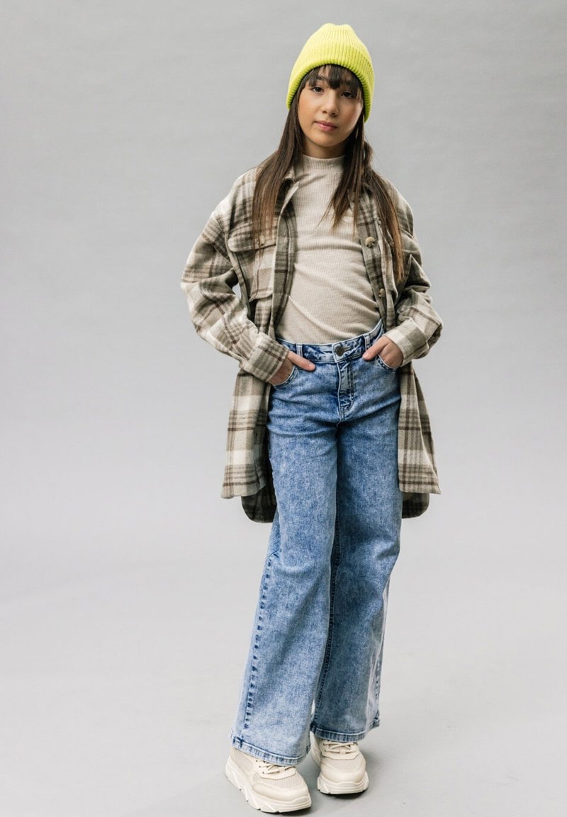 Bruine geruite shirtjacket over een beige top, blauwe wijde jeans en crèmekleurige sneakers. Felgele gebreide beanie. Grauwe achtergrond.