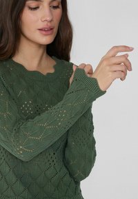 Grüner Strickpullover mit einem gewellten Ausschnitt und einem Rautenmuster mit offenen Maschen an den Ärmeln und dem Körper.