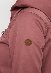 Sako barvy prachové růže s hladkou texturou, vybavené kapsou se zipem a hnědou koženou nášivkou s logem na rameni. Navrženo s kapucí.