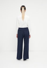 Witte overhemd met kraag, gecombineerd met marineblauwe broek met pinstripe en wijde pijpen. De broek heeft een elastische tailleband en achterzakken. Persoon staat met de rug naar de kijker.