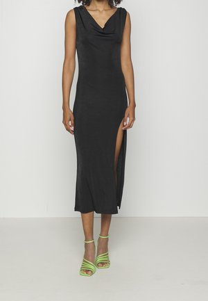 Kobieta ubrana w czarną midi sukienkę bez rękawów z bocznym rozcięciem oraz neonowo-zielone sandały na wysokim obcasie ze strapsami, stojąca na tle jednolitego białego tła.