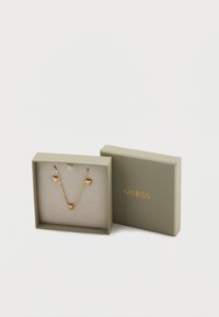 Χρυσό κολιέ σε σχήμα καρδιάς, τοποθετημένο σε πράσινο κουτί με μαλακό εσωτερικό. Το καπάκι φέρει ανάγλυφο λογότυπο. Το κουτί έχει καθαρό, κομψό σχεδιασμό.