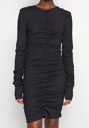 Osoba ubrana w dopasowaną czarną sukienkę z długim rękawem, ozdobioną białymi pionowymi prążkami oraz marszczeniami z przodu i na rękawach.