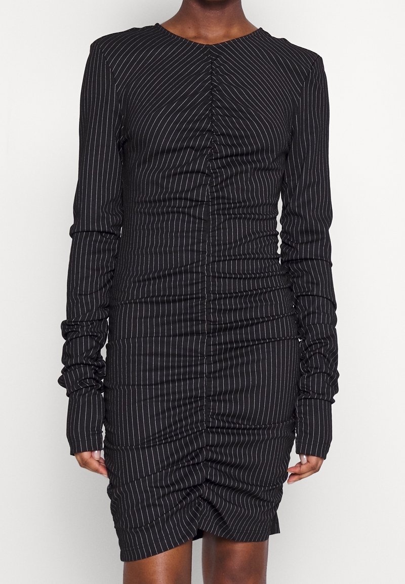 Personne portant une robe noire à manches longues ajustée avec des fines rayures blanches verticales et un effet froncé au centre devant et sur les manches.