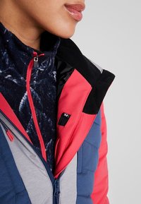 Bunte Jacke mit roten, blauen und grauen Paneelen und einem gesteppten Design. Enthält einen marineblauen Fleece-Kragen und eine vordere Reißverschlussöffnung mit einem Logo-Tag.