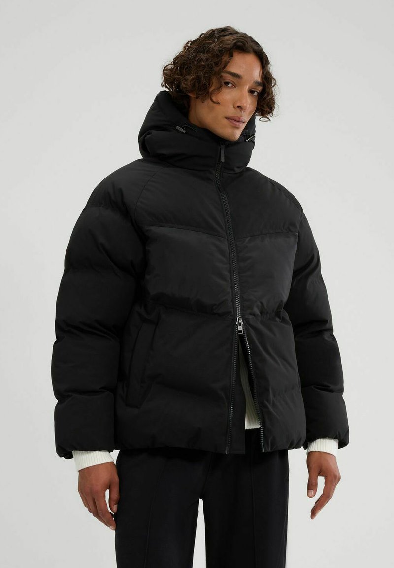 Chaqueta de plumas negra con cuello alto y cierre de cremallera. Presenta paneles acolchados, una silueta cuadrada y puños elásticos. Textura suave y mate.