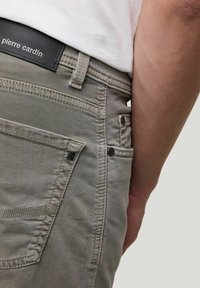 Graue Baumwollhose mit sichtbaren Nähten, seitlichen Taschen und einem schwarzen Lederlabel mit "pierre cardin" am Bund.