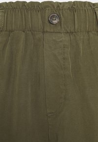 Gros plan sur la ceinture d'un pantalon vert olive avec un haut élastique, un bouton visible unique et des coutures détaillées le long de la braguette.
